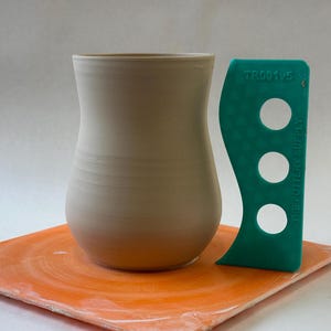 May include: A light beige ceramic vase with a rounded bottom and a narrow opening. The vase is sitting on an orange pottery wheel.