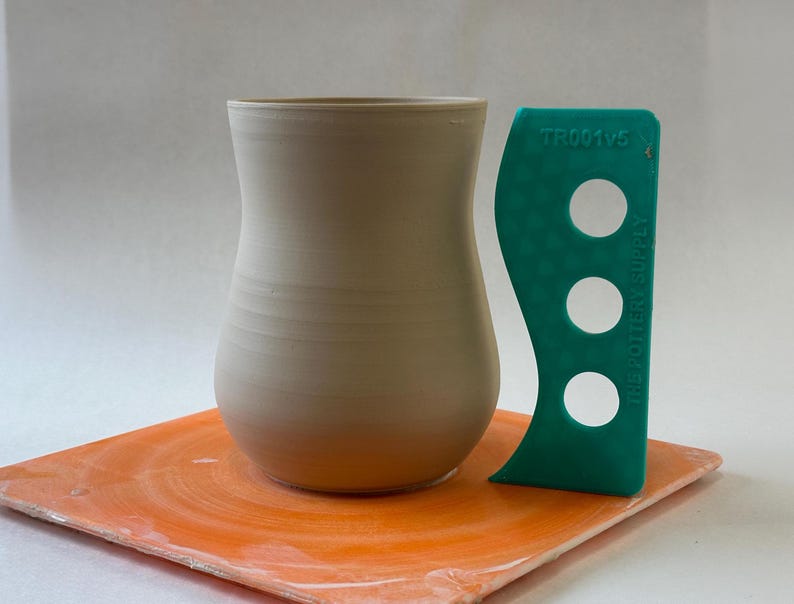May include: A light beige ceramic vase with a rounded bottom and a narrow opening. The vase is sitting on an orange pottery wheel.