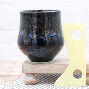 Puede incluir: Una taza de cerámica azul oscuro con un esmalte marrón y un efecto de goteo. La taza está sentada en un soporte de madera con una herramienta amarilla al lado.