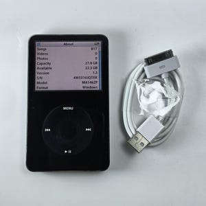 Może przedstawiać: Czarny iPod z białym kablem i ładowarką. Ekran wyświetla informacje o urządzeniu, w tym liczbę utworów, pojemność i dostępną przestrzeń. iPod posiada pokrętło i przyciski sterujące.