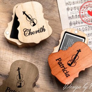 Könnte beinhalten: Zwei hölzerne Geigenförmige Boxen mit Deckeln. Die obere Box ist hellbraun mit dem Namen "Cherith" eingraviert. Die untere Box ist dunkelbraun mit dem Namen "Patricia" eingraviert. Beide Boxen haben ein Geigenmotiv auf dem Deckel eingraviert.