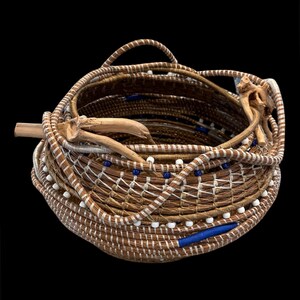 May include: A round, hand-coiled basket constructed from brown natural fibers, featuring white and blue accent beads woven into the structure. The rim is adorned with a piece of light-colored driftwood, and the exterior displays a cross-stitch pattern.
