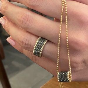 May include: A gold chain necklace with a grey and gold beaded cylinder pendant. The necklace is worn by a person with a grey and gold beaded ring on their finger.