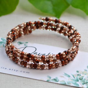 May include: A spiral wrap bracelet with three strands of small, round beads in shades of brown, copper, and clear. The bracelet is resting on a white card with the Etsy shop name "Davalia" printed on it.