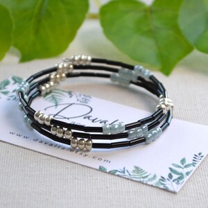 Peut inclure: Un bracelet à plusieurs brins avec un cordon noir, des perles argentées et des perles bleu clair. Le bracelet est présenté sur une carte blanche avec un motif floral et l'adresse du site web www.davaliaetsy.com.