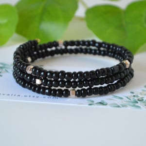 May include: A multi-strand wrap bracelet composed of small, glossy black seed beads. The design features evenly spaced, metallic silver-toned hexagonal accent beads integrated throughout the three circular rows of the flexible, coiled wire structure.