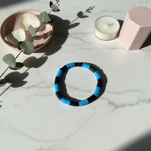 May include: A circular bracelet composed of alternating flat, disc-shaped beads in vibrant cyan blue and solid black. The beads are arranged in a repeating pattern, creating a simple, uniform band that rests on a smooth, white marble surface.