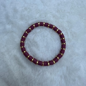 Puede incluir: Una pulsera circular compuesta por una alternancia de cuentas de disco de color ciruela oscuro y pequeñas cuentas espaciadoras metálicas de tono dorado reflectante. Las cuentas están dispuestas en un patrón repetitivo y uniforme.