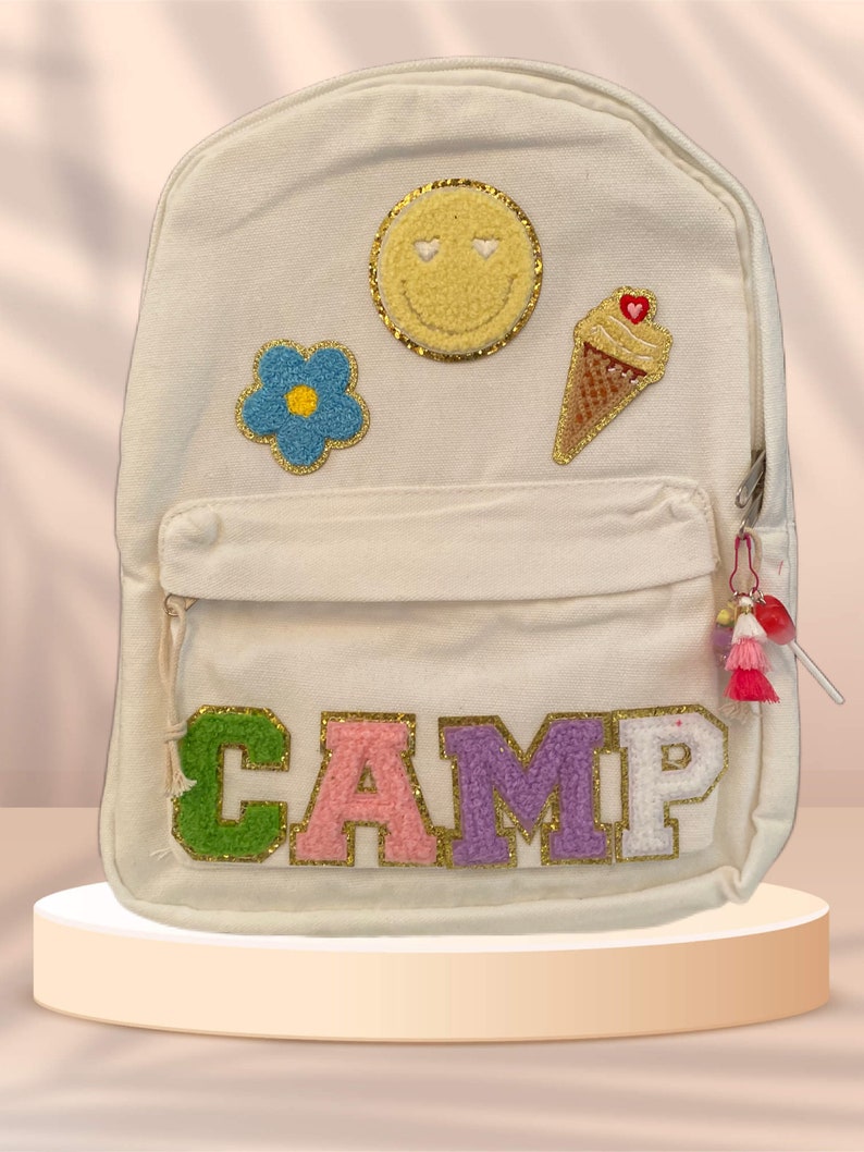 Puede incluir: Mochila de lona blanca con un "CAMP" bordado en oro en la parte delantera. La mochila tiene varios parches coloridos, incluyendo una cara sonriente, una flor y un cono de helado.