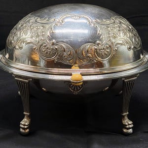 Könnte beinhalten: Versilberte Servierschüssel mit gewölbtem Deckel und aufwendigen floralen Details. Die Schüssel hat einen runden Boden und wird von vier dekorativen Beinen getragen. Der Deckel hat einen zentralen Griff und ist angelenkt. Ein klassisches Stück zum Servieren von Speisen.