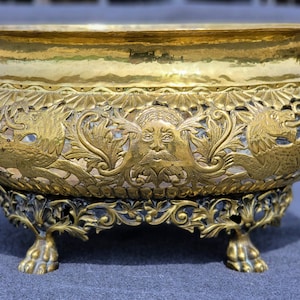 Puede incluir: Un macetero de metal dorado con intrincados diseños florales y animales. El macetero tiene tres patas y una base redonda.