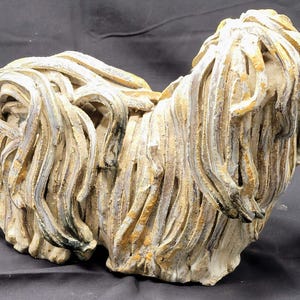 May include: A ceramic sculpture of a dog, featuring a textured, layered design. The dog's fur is depicted with long, flowing strands in shades of beige, cream, and hints of gray and yellow. The sculpture has a weathered appearance.