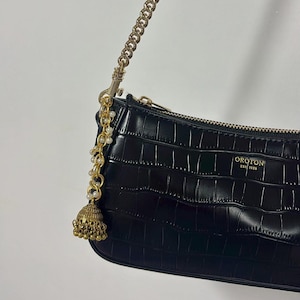 Könnte beinhalten: Eine schwarze Handtasche mit Krokodilprägung und einem goldfarbenen Kettenriemen. Die Tasche trägt den Schriftzug OROTON EST 1938 in Gold. Ein goldfarbener Jhumka-Anhänger mit kleinen weißen Perlen ist am Kettenriemen der Tasche befestigt.
