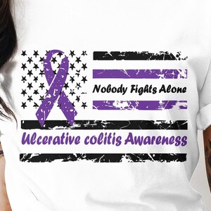 May include: White t-shirt with a purple ribbon and a distressed American flag design. The text "Nobody Fights Alone" and "Ulcerative Colitis Awareness" is printed on the shirt.