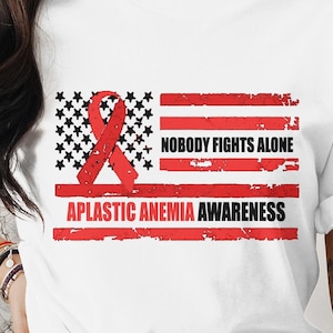 Può includere: Maglietta bianca con un nastro rosso e un design a bandiera americana. Il testo "Nobody Fights Alone" e "Aplastic Anemia Awareness" è stampato sulla maglietta.