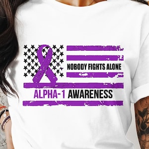 May include: White t-shirt with a purple ribbon and an American flag design. The flag has the words "Nobody Fights Alone" and "ALPHA-1 AWARENESS" printed on it.