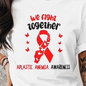 Può includere: Maglietta bianca con il testo "We fight together" e un nastro rosso con farfalle. Il testo "Aplastic Anemia Awareness" è sotto il nastro.