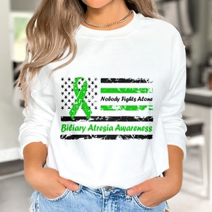 Op de afbeelding: Witte longsleeve met een groen lint en een verweerd Amerikaans vlagontwerp. De tekst "Nobody Fights Alone" en "Biliary Atresia Awareness" staat op de trui gedrukt.
