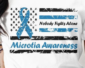 May include: A white t-shirt with a blue ribbon and a distressed American flag design. The text "Nobody Fights Alone" and "Microtia Awareness" is printed on the shirt.
