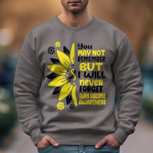 Op de afbeelding: Grijs sweatshirt met een gele zonnebloem en zwarte tekst die "You may not remember but I will never forget Ewing Sarcoma Awareness" luidt.