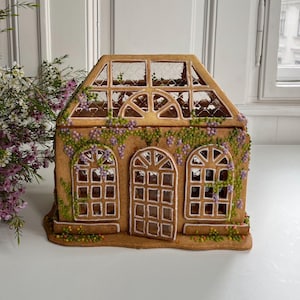 Op de afbeelding: Een gingerbreadhuis in de vorm van een kas met een bruin dak, witte versieringen en een doorzichtig plastic dak. Het huis is versierd met paarse en groene glazuur om eruit te zien als wijnstokken en bloemen.