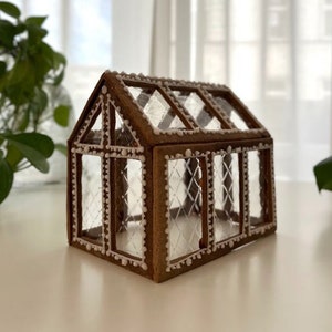 Könnte beinhalten: Ein Lebkuchenhaus mit brauner Außenseite und weißen Zuckergussdetails. Das Haus hat klare, rautenförmige Fenster und ein Satteldach. Die Ränder sind mit kleinen weißen Punkten verziert, was an ein Gewächshaus erinnert.