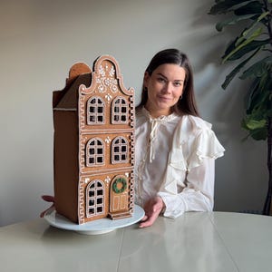 May include: A tall, three-story gingerbread house structure featuring brown baked walls, white icing detailing around the arched windows, and a decorative white icing pattern on the roof gable. A small green wreath is attached to the front door.