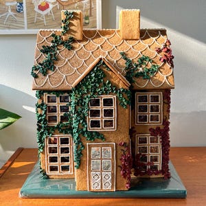 Può includere: Una casetta di pan di zenzero decorata con glassa verde e rossa, che ricorda l'edera che cresce sui lati. La casa ha un tetto di glassa bianca con un motivo a festoni.