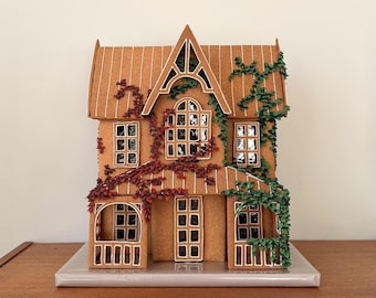 May include: A gingerbread house model featuring a brown exterior with white piped trim around the windows, doors, and roofline. The structure is decorated with red and green icing vines climbing the walls and roof, set on a silver base.