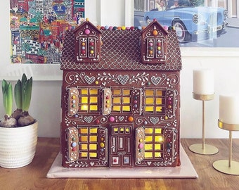 May include: A gingerbread house decorated with white icing, colorful candies, and lights. The house has a brown roof, white trim, and windows with shutters. The house is sitting on a pink surface.