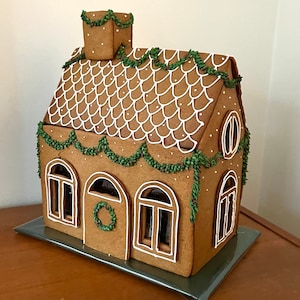 Op de afbeelding: Een gingerbreadhuis versierd met groene en witte glazuur. Het dak is bedekt met witte glazuur en heeft een patroon van kleine witte stippen. Het huis heeft vier ramen en een deur. Er is een krans op de deur.