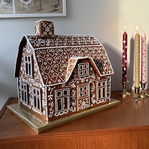 Può includere: Una casetta di pan di zenzero decorata con glassa bianca e cuori, appoggiata su un vassoio dorato. Tre candele con pois rossi, rosa e bianchi sono sullo sfondo.