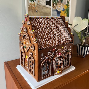 Op de afbeelding: Een gingerbreadhuis versierd met kleurrijke snoepjes en witte glazuur. Het huis heeft een bruin dak met witte gegolfde randen en een wit hek.