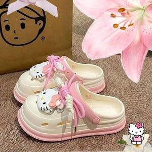 Op de afbeelding: Witte en roze platformklompen met een Hello Kitty-ontwerp. De klompen hebben een roze rand en veters, met een Hello Kitty-gezicht aan de bovenkant. Een roze strik en een roze bloem zijn ook zichtbaar.