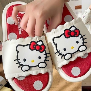 Op de afbeelding: Witte en rode Hello Kitty-slippers. De slippers hebben een wit bovenwerk met een Hello Kitty-gezicht en een rode strik. Het voetbed is rood met witte stippen. De zool is wit.