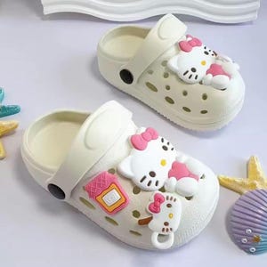Op de afbeelding: Twee witte kinderschoenen met Hello Kitty-versieringen. Elke schoen heeft een witte band en is versierd met roze strikken en cartoonkarakterontwerpen. De schoenen hebben een geperforeerd ontwerp.
