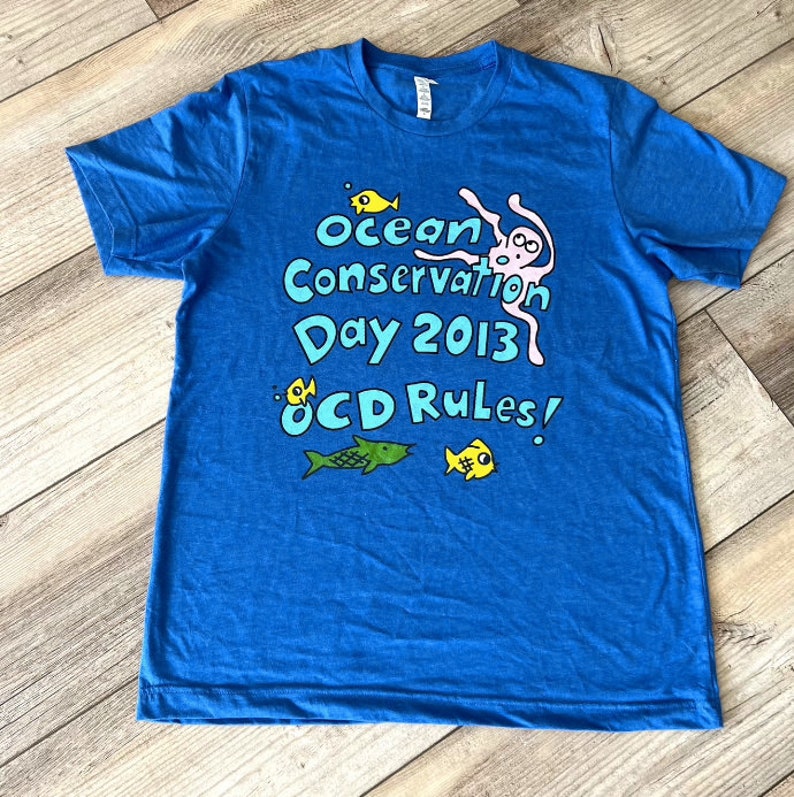 May include: A blue t-shirt with a graphic design of an octopus, fish, and the text "Ocean Conservation Day 2013 OCD Rules!"
