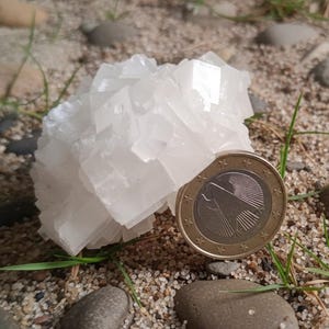 May include: A cluster of white, crystalline mineral formations, possibly a geode or crystal, next to a one Euro coin. The coin is gold-coloured with a silver centre. The mineral is on a bed of sand, small rocks, and grass.