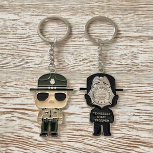 May include: Two silver keychains with cartoon figures. One keychain features a cartoon police officer with a green hat and sunglasses. The other keychain features a black and silver badge with the text "Tennessee Highway Patrol" and "Trooper".
