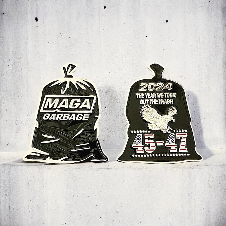 May include: Two black and gold garbage bag shaped pins. The first pin says "Maga Garbage". The second pin says "2024 The Year We Took Out The Trash 45-47".