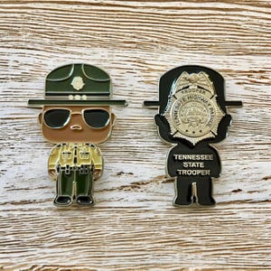 May include: Two enamel pins featuring Tennessee State Trooper designs. One pin depicts a trooper in uniform with sunglasses and a hat. The other pin shows a badge with the text "Tennessee State Trooper".