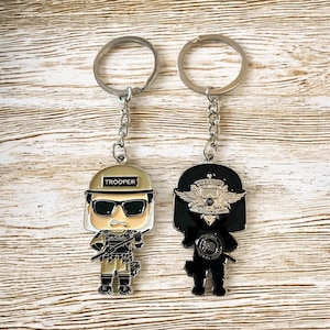 May include: Two metal keychains featuring stylized figures. One keychain depicts a trooper with the word "TROOPER" on the helmet. The other keychain shows a figure with a badge and wings. Both keychains have silver key rings and chains.