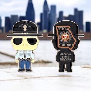 May include: Two enamel pins featuring a Georgia State Trooper design. One pin depicts a trooper in uniform with sunglasses, the other shows the state outline with text: "Georgia State Patrol" and "Georgia State Trooper".
