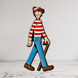 Op de afbeelding: Een kleurrijke emaille pin met een stripfiguur in een rood-wit gestreept shirt, een blauwe spijkerbroek, bruine schoenen en een bijpassende hoed. Het personage houdt een bruine wandelstok vast.