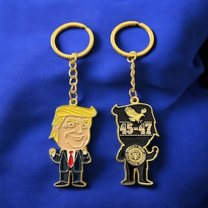 May include: Two gold keychains with cartoon figures. One keychain features a caricature of Donald Trump in a blue suit and red tie. The other keychain features a caricature of Donald Trump in a black suit with the text "45-47" on the chest.
