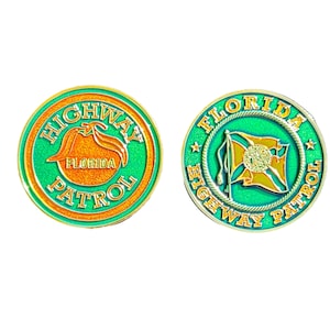 Op de afbeelding: Twee goudkleurige challenge-munten met groene en oranje accenten. De voorkant van de munt heeft de woorden "HIGHWAY PATROL" met een ontwerp van de vlag van de staat Florida. De achterkant van de munt heeft de woorden "FLORIDA HIGHWAY PATROL" met een touwontwerp.