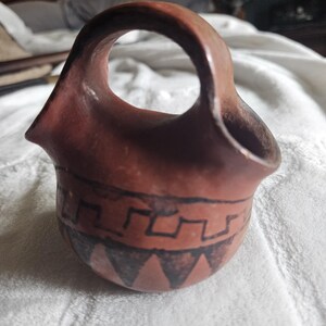 May include: A small, hand-crafted, terracotta-colored ceramic vessel with a handle. The vessel features a geometric design of black lines and triangles around the base. The opening is circular, and the handle is integrated into the design.