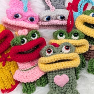 May include: A collection of handmade crochet frog-shaped items in various colors. The frogs have large, expressive eyes and open mouths. Some have pink or yellow accents, and one has a pink heart.