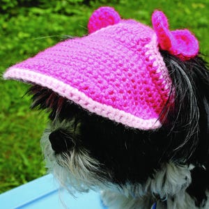 May include: A pink crocheted visor with a light pink trim and two pink ear-shaped accents. The visor is worn by a small dog with black and white fur. The dog is sitting outside on a blue surface.