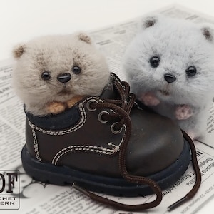 Puede incluir: Dos juguetes de animales de crochet, uno gris y otro beige, llevando una bota de cuero marrón. El juguete gris es un hámster o un ratón, y el juguete beige es un oso o un conejo. La bota está sobre un fondo de periódico.